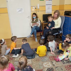 Zdjęcie przedstawia kobietę czytającą dzieciom książkę, po prawej znajduje się kobieta z dzieckiem, które siedzi jej na kolanach. Wokół nich siedzą dzieci odwrócone tyłem, patrzące w stronę kobiet.