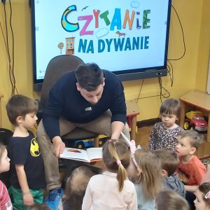 Zdjęcie przedstawia mężczyznę ubranego w ciemne ubrania, nachylinego nad dziećmi, czytającego książkę dzieciom, które są skupione wokół niego. Za plecami widoczny jest napis na tablicy "Czytanie na dywanie".