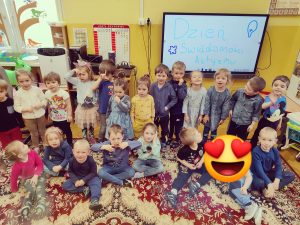 Zdjęcie przedstawia grupę dzieci. Większość dzieci ma na sobie niebieski element garderoby. Za nimi na tablicy widnieje napis "Dzień Świadomości Autyzmu"