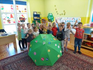 Zdjęcie przedstawia dzieci, które stoją za zieloną parasolką, na której znajdują się kolorowe kropki.