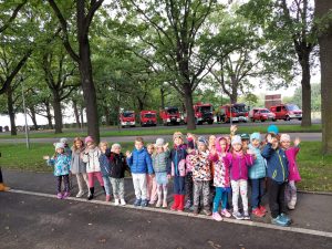 Zdjęcie przedstawia dzieci stojące wzdłuż linii, namalowanej na asfalcie Za nimi widać drzewa i wozy strażackie.