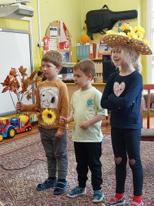 Zdjęcie przedstawia dwóch chłopców i dziewczynkę. Chłopiec trzyma w ręce słonecznik, dziewczynka ma kapelusz na głowie.