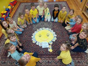 Zdjęcie przedstawia słonecznik z papieru położony na dywanie, wokół którego siedzą dzieci ubrane na zółto.